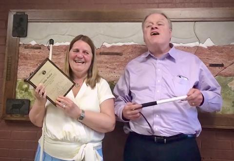 Julie Deden, smiling, holds up a plaque commemorating 25 years of Service at the Colorado Center for the Blind, presented by a smiling Gary Wunder from the Board of Directors.